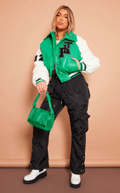Person wearing a green and white varsity jacket with black pants, holding a green handbag against a beige background.