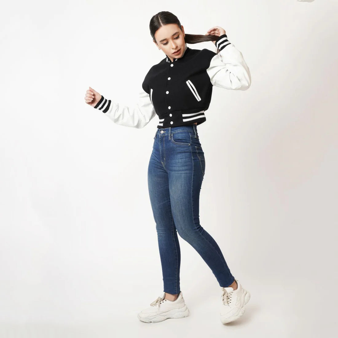 Woman wearing a black and white varsity jacket, blue jeans, and white sneakers on a white background