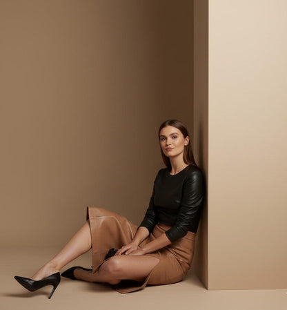 Woman sitting against a beige wall wearing a black top and brown skirt.
