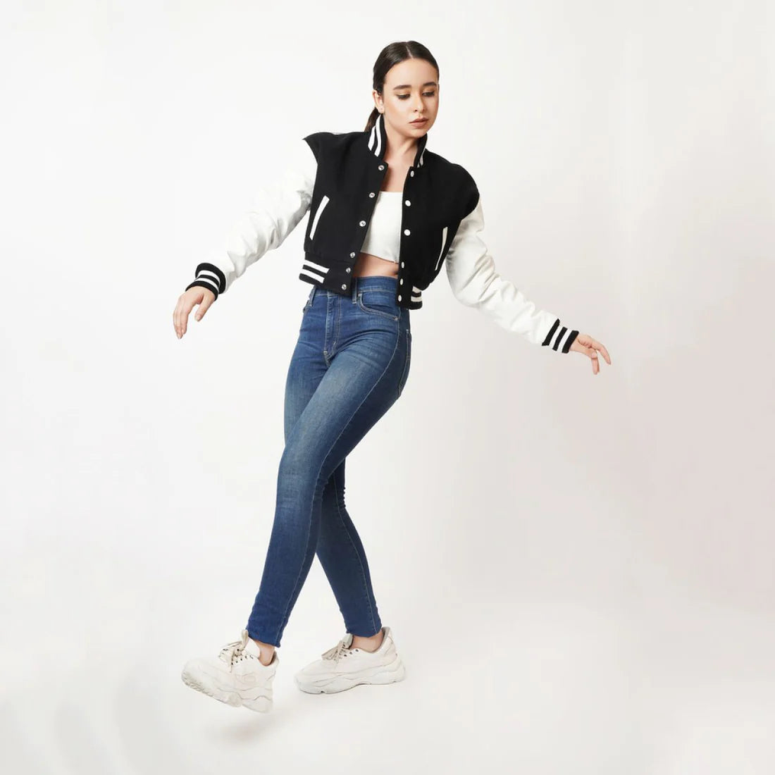Person wearing a black and white varsity jacket, blue jeans, and white sneakers on a white background