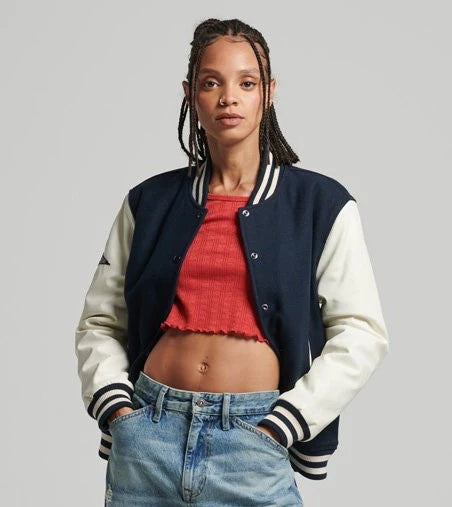 Woman wearing a navy and white varsity jacket over a red top and blue jeans on a gray background