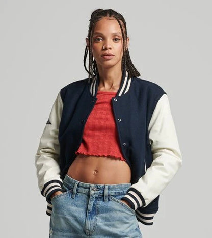 Woman wearing a navy and white varsity jacket over a red top and blue jeans on a gray background