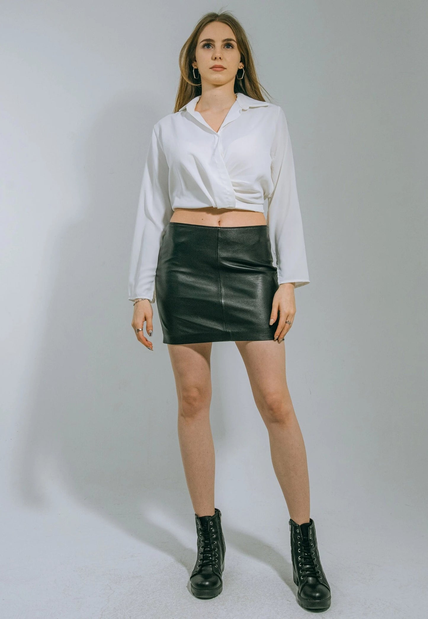 Woman wearing a white blouse, black leather skirt, and black boots on a plain background