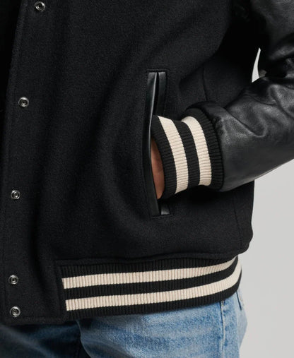 Black varsity jacket with white stripes on cuffs and waistband, worn over a leather jacket.